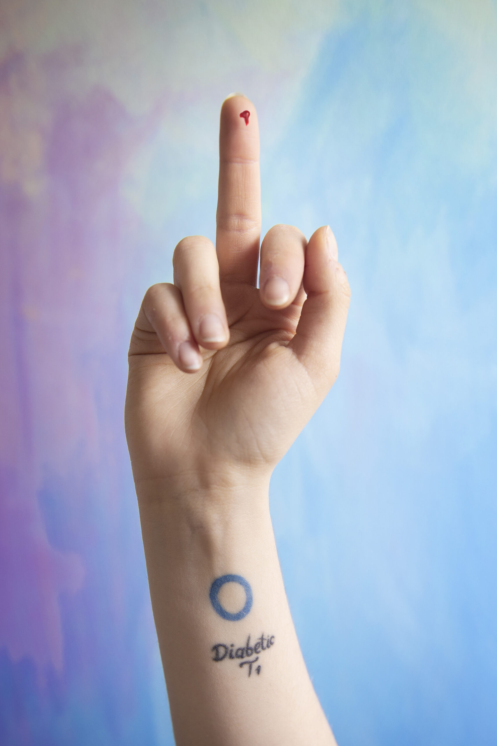 Zdjęcie przedstawia dłoń ukazującą środkowy palec. Na ręce znajduje się tatuaż z napisem cukrzyca po angielsku. Z palca cieknie krew jak przy badaniu poziomu cukru we krwi. Tło zdjęcia jest kolorowe, wesołe i kontrastuje z wymownym, wulgarnym znakiem środkowego palca.